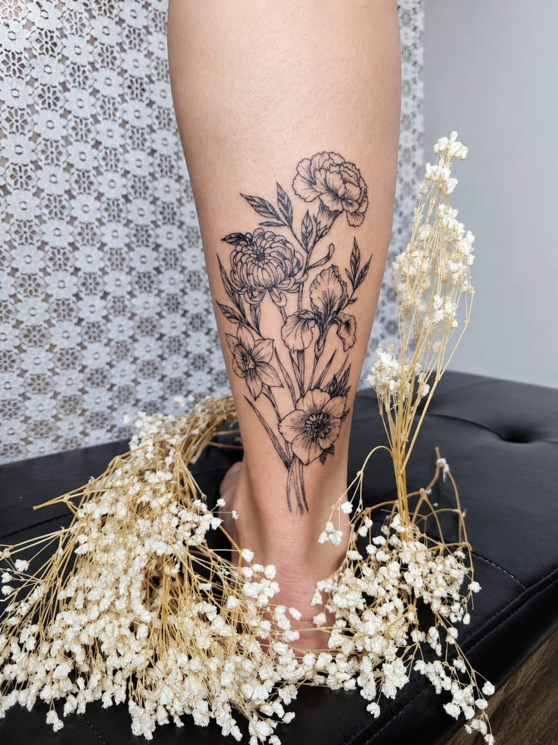 Bouquet de fleurs en trait fin sur le mollet : œillet, chrysanthème, iris, narcisse et une fleur ouverte, entouré de gypsophile séchée.