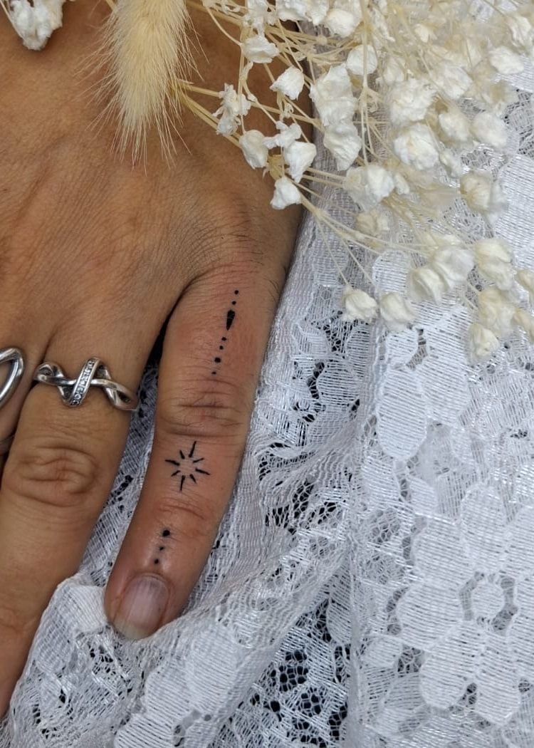 Petite étoile et lignes de points tatouées sur le côté d’un doigt, main posée sur de la dentelle avec de la gypsophile séchée.