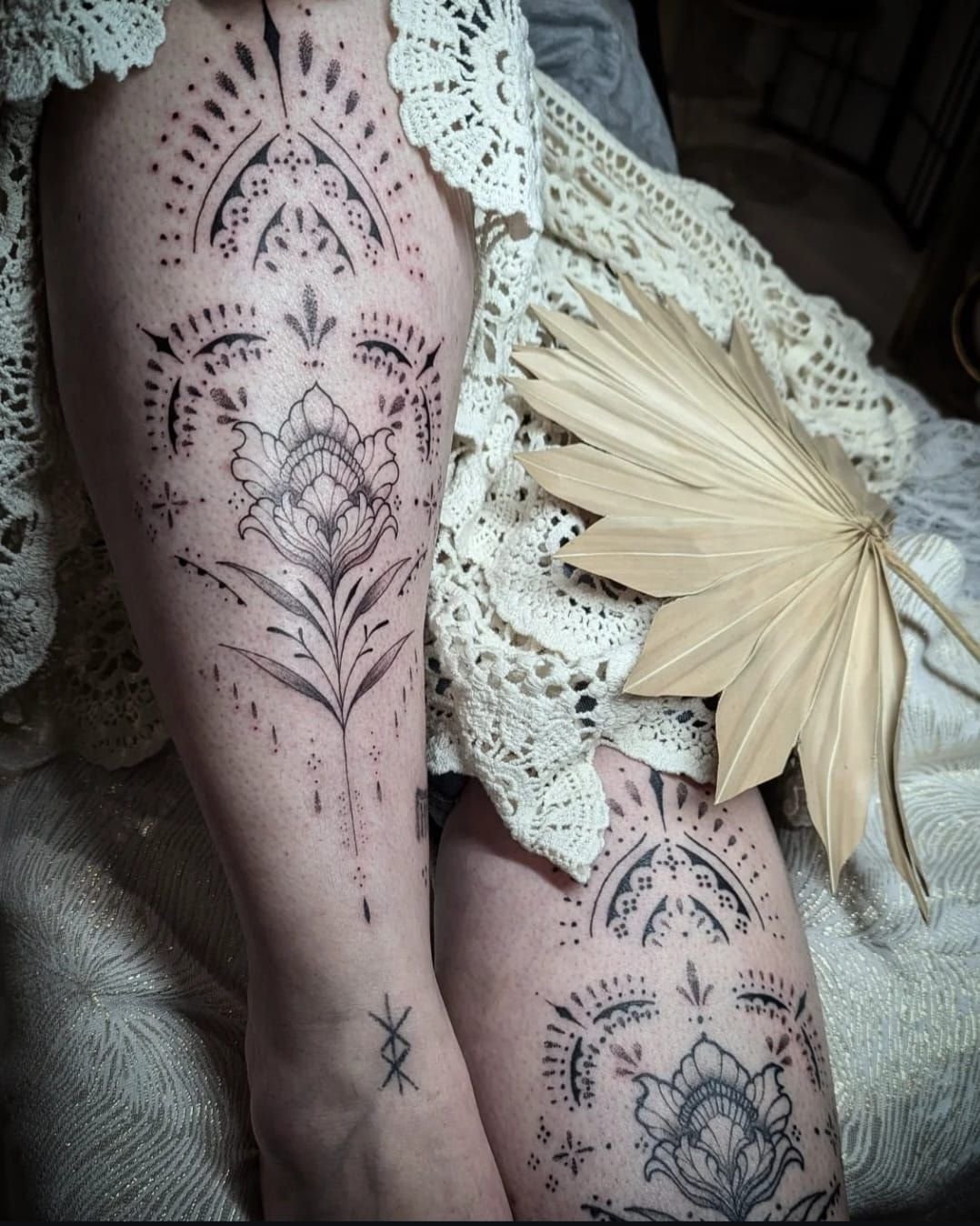 Deux tatouages ornementaux assortis en trait fin et en points : arcs de dentelle pointillée et fleur stylisée, posés sur un tissu en crochet avec une palme séchée.