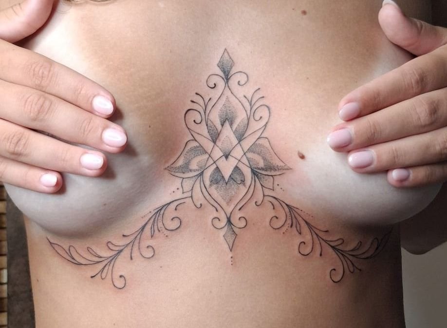 Tatouage ornemental sous la poitrine : motif central de losanges et de pétales ombrés en points, prolongé de volutes et de feuillages fins de chaque côté.