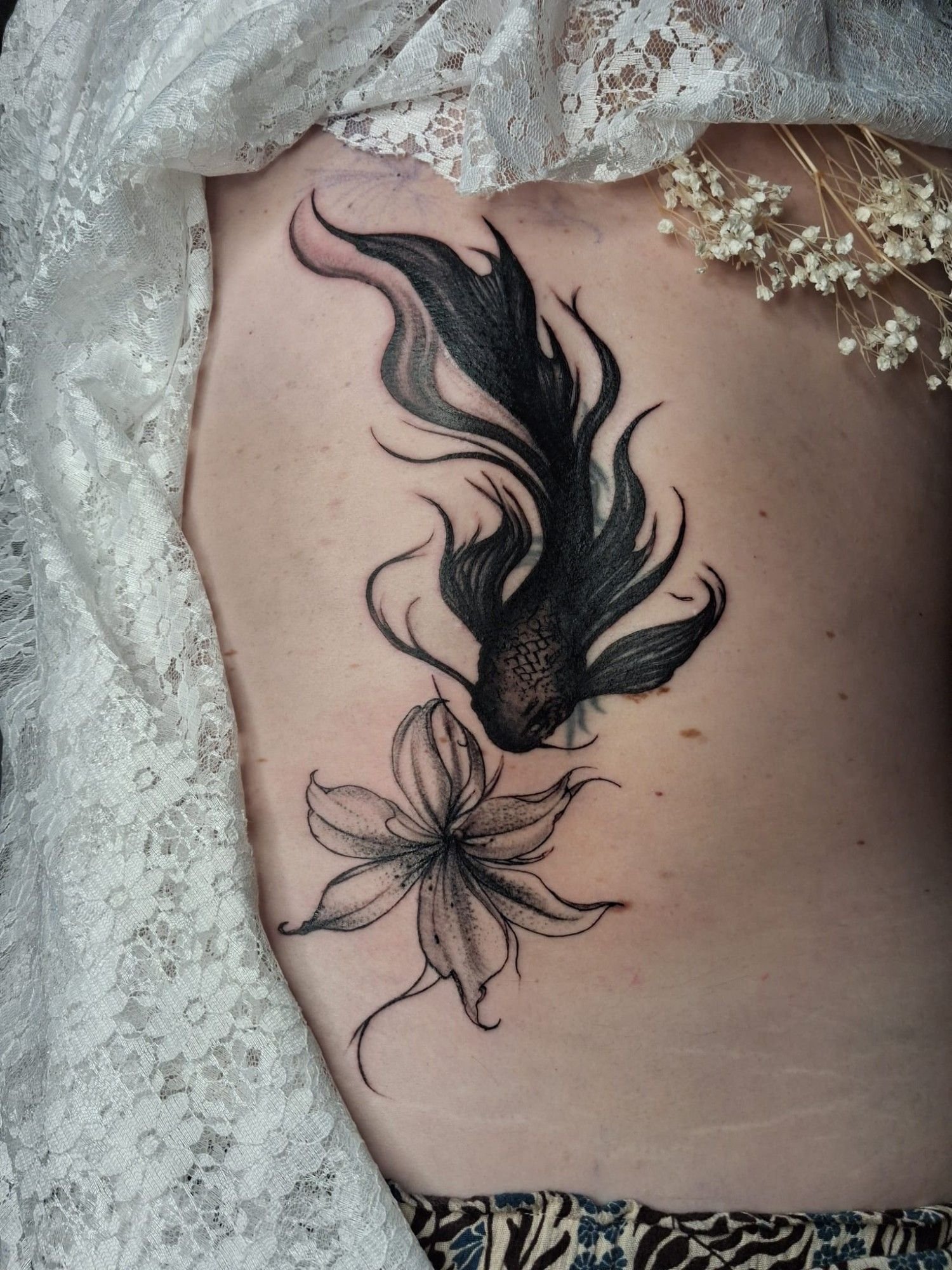 Poisson aux longues nageoires voilées en black and grey, au-dessus d’une fleur de lys en trait fin, sur le flanc : un cover qui recouvre un ancien tatouage.