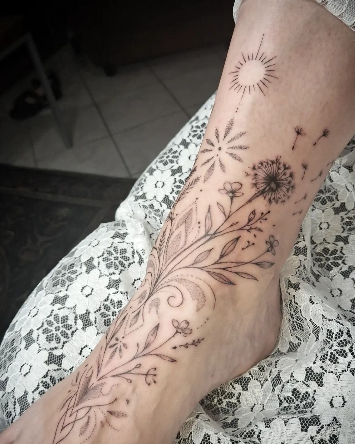 Composition fine line sur la cheville et le pied : un soleil rayonnant, un pissenlit dont les aigrettes s’envolent, des fleurs, des feuillages et des ornements en points.