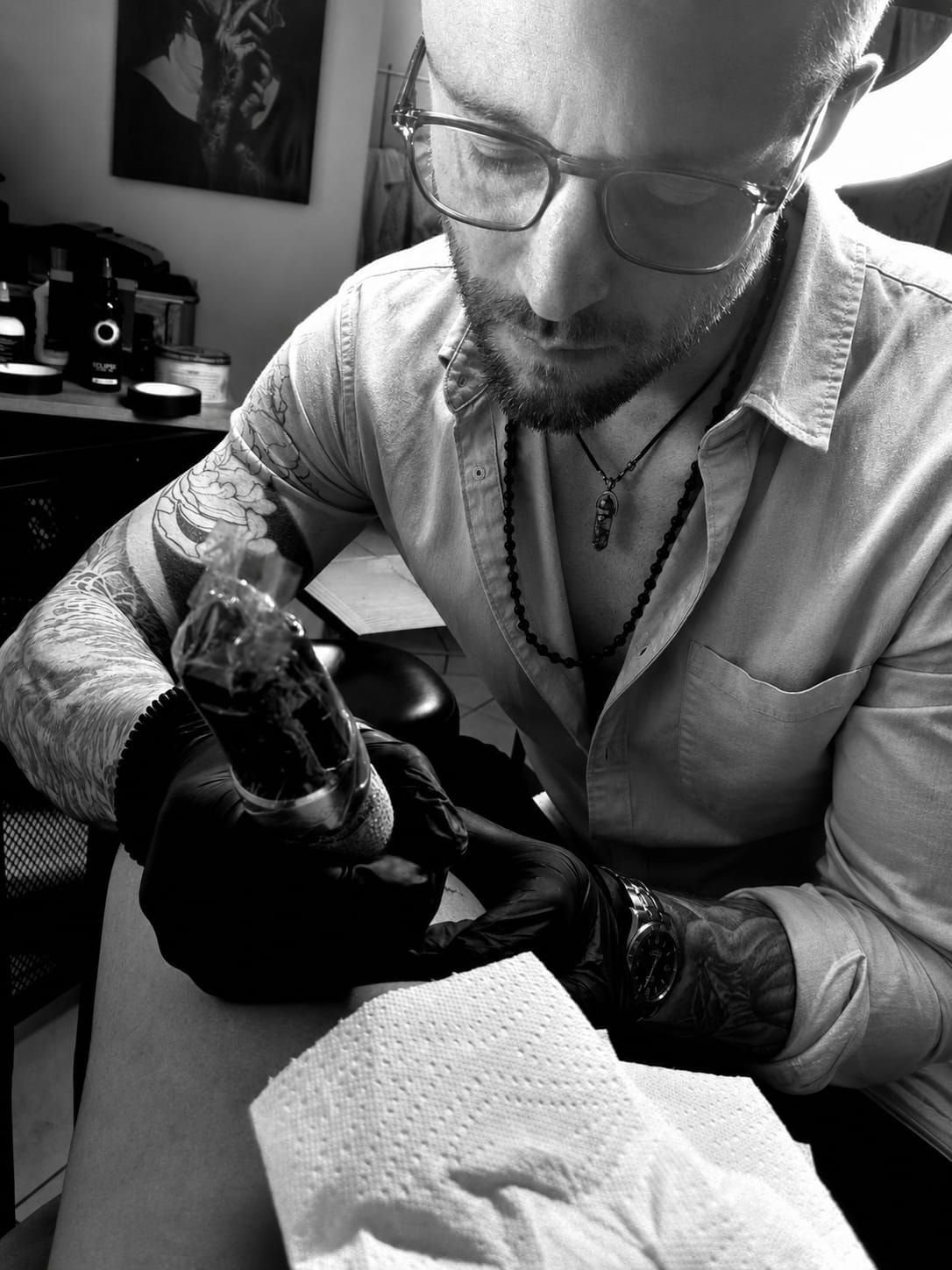 Tidjé, lunettes et chemise claire, penché sur un tatouage en cours, gants noirs et machine en main.